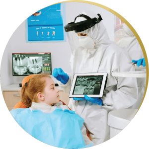 A dentist in full protective gear shows a child their dental X-ray on a tablet.