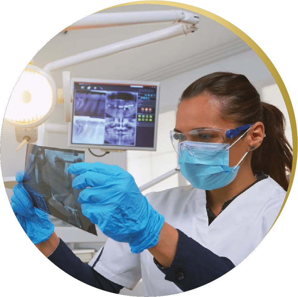 A female dentist wearing a mask, safety glasses, and blue gloves is examining a dental X-ray. A computer monitor in the background displays additional dental scans